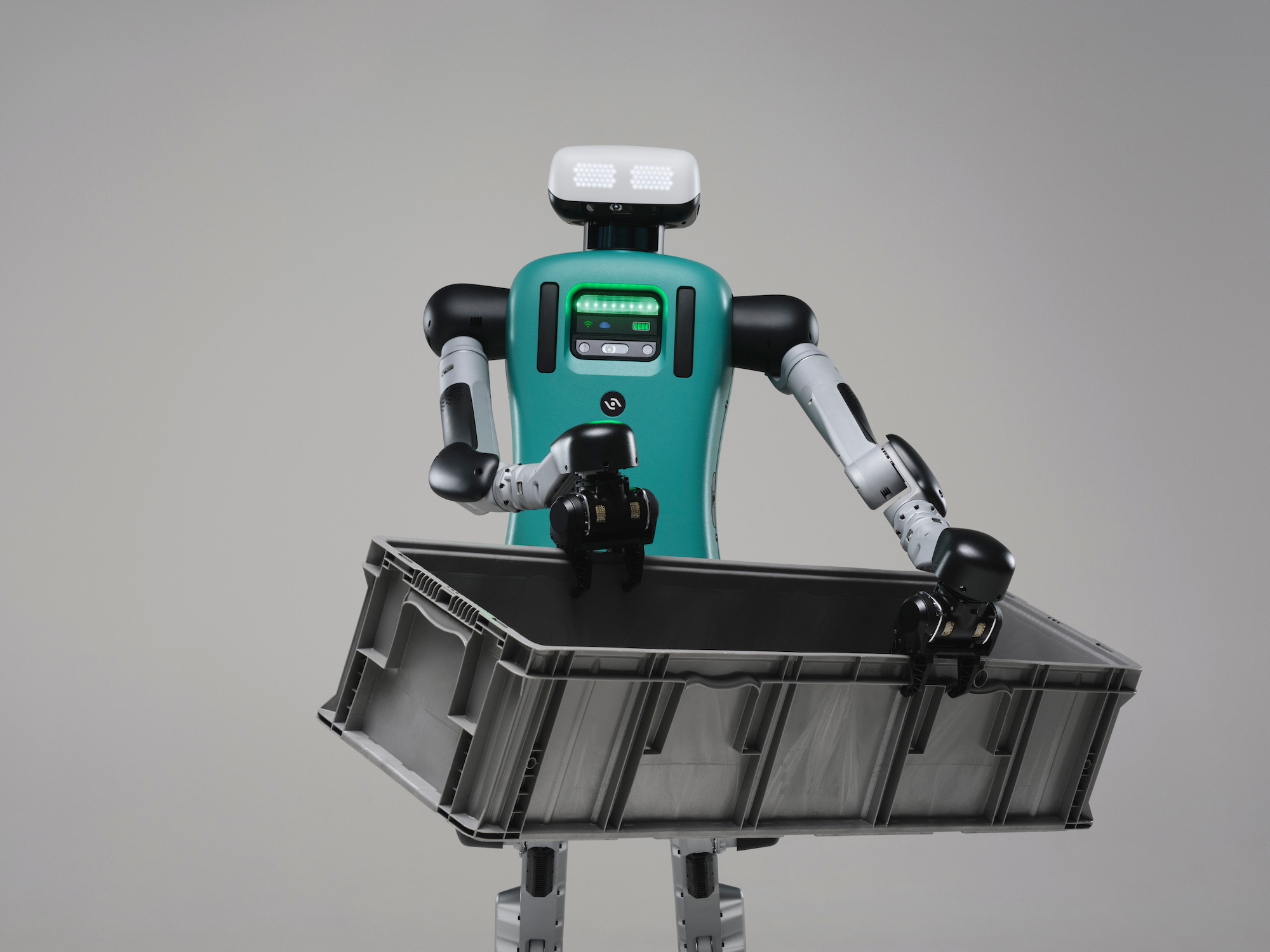An earlier-generation Agility Digit humanoid holds a grey plastic tote in a studio photograph.